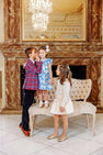 Three children pose indoors on an elegant cream-colored couch. A boy kisses a girl’s head while another girl looks on. The girls are dressed up for the holidays in The Beaufort Bonnet Company’s Grandccasion™ Delaney Dinner Dress - Bagley Bows (Beale Street Blue), all gathered in front of a large mirror and chandelier.