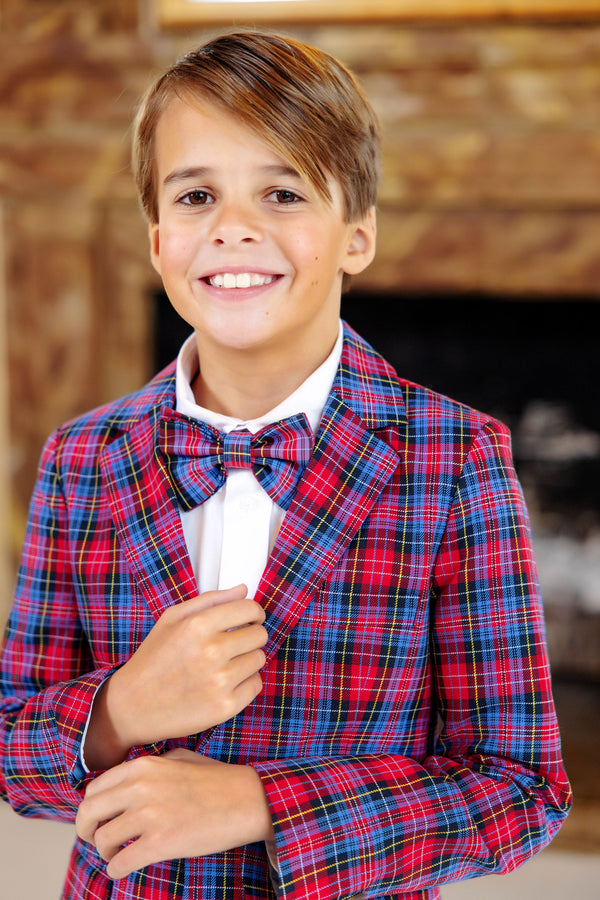 A smiling young boy stands indoors wearing a white shirt and a Baylor Bowtie - Bucktown Tartan by The Beaufort Bonnet Company, paired with a red and blue plaid suit.