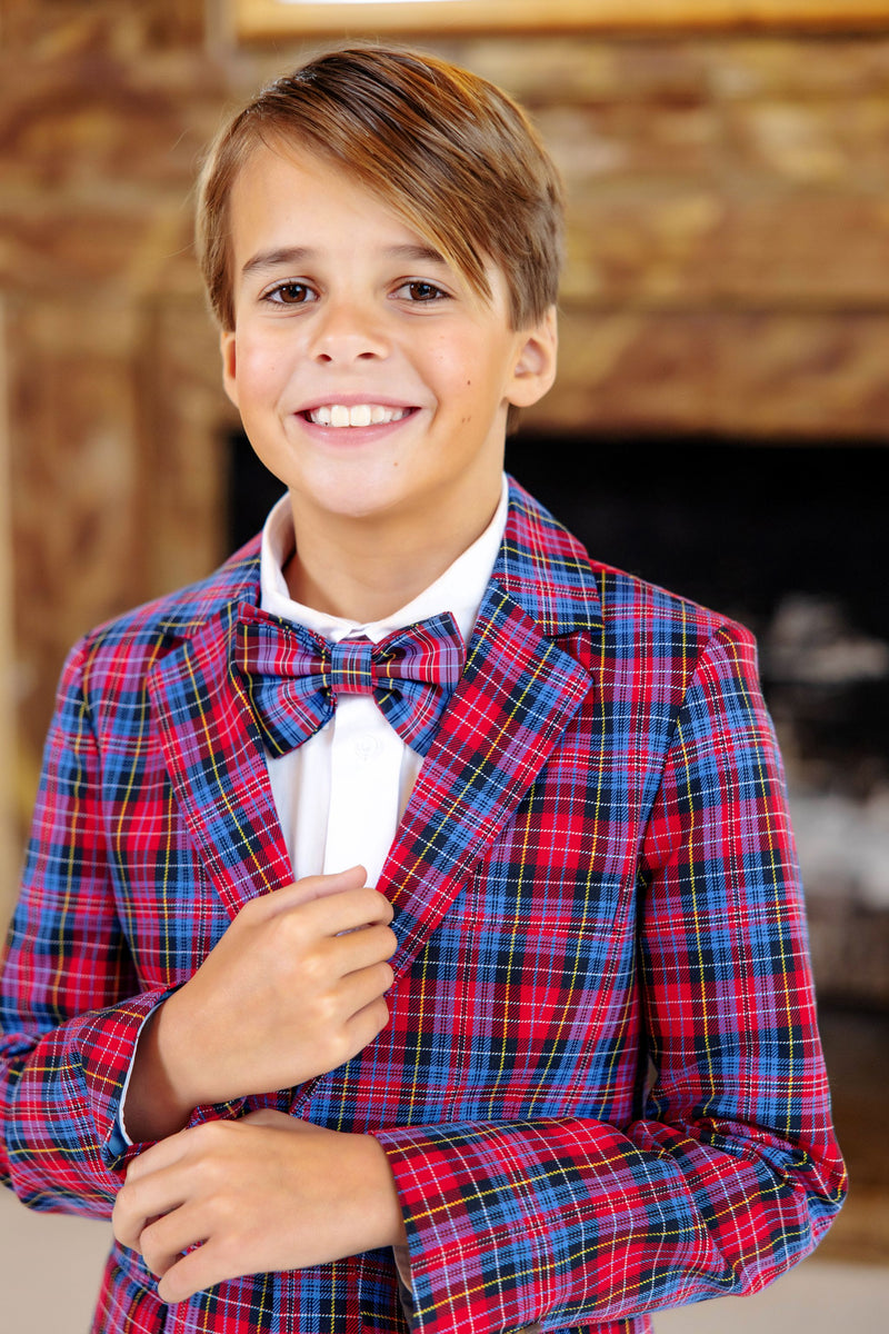 A smiling young boy stands indoors wearing a white shirt and a Baylor Bowtie - Bucktown Tartan by The Beaufort Bonnet Company, paired with a red and blue plaid suit.