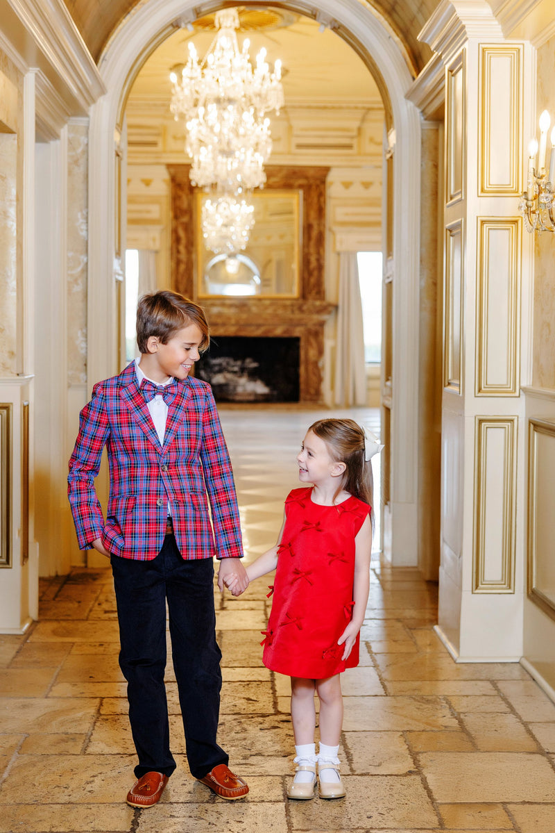 A boy dressed in The Beaufort Bonnet Company’s Prep School Pants – Newport Night Corduroy with Richmond Red Stork, a plaid blazer, and a bow tie holds hands with a girl in a red dress. They smile at each other in an elegant hallway adorned with chandeliers and ornate decor.