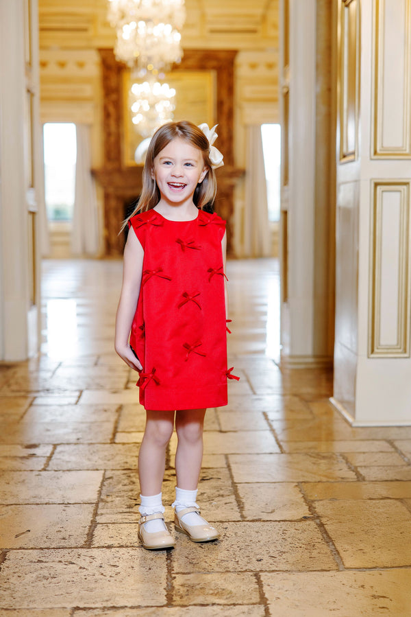 A young girl stands smiling on a tiled floor in an elegant, ornate room with chandeliers and gold accents, wearing The Beaufort Bonnet Company’s Grandccasion™ Jilly Jumper in Richmond Red Taffeta adorned with bows.