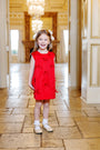 A young girl stands smiling on a tiled floor in an elegant, ornate room with chandeliers and gold accents, wearing The Beaufort Bonnet Company’s Grandccasion™ Jilly Jumper in Richmond Red Taffeta adorned with bows.