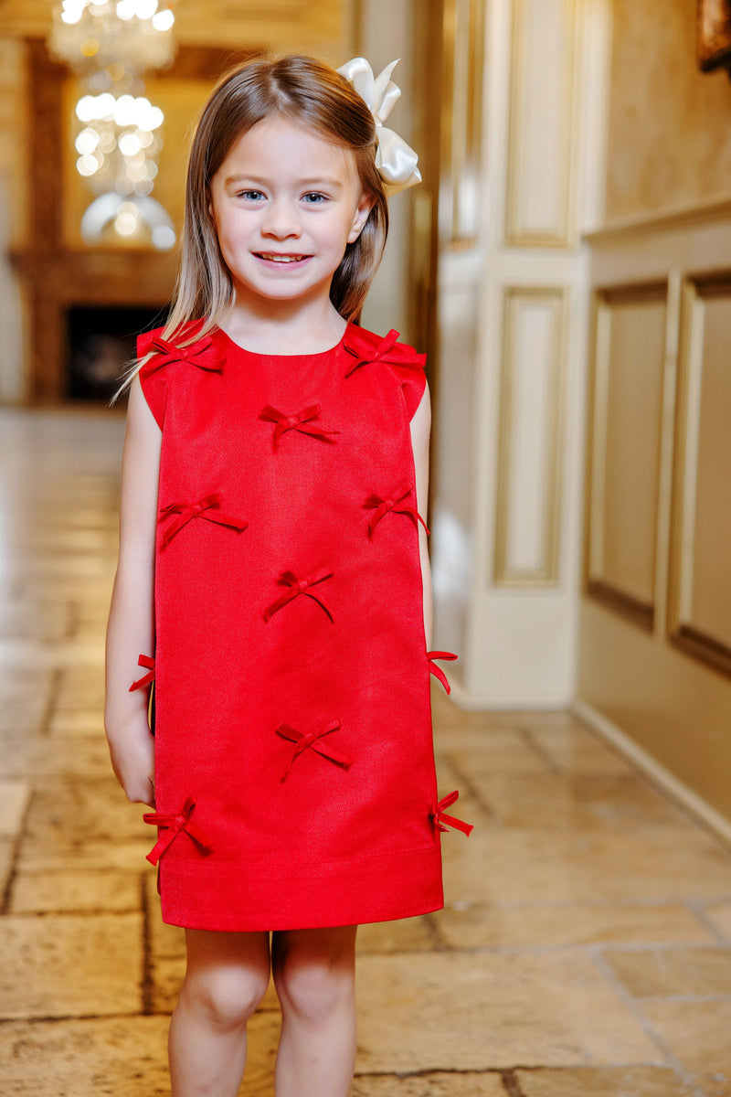 A young girl with long brown hair and a white bow wears The Beaufort Bonnet Company’s Grandccasion™ Jilly Jumper in Richmond Red Taffeta, which features charming bow accents. She stands indoors on a tiled floor, smiling at the camera, with an elegant blurred hallway in the background.