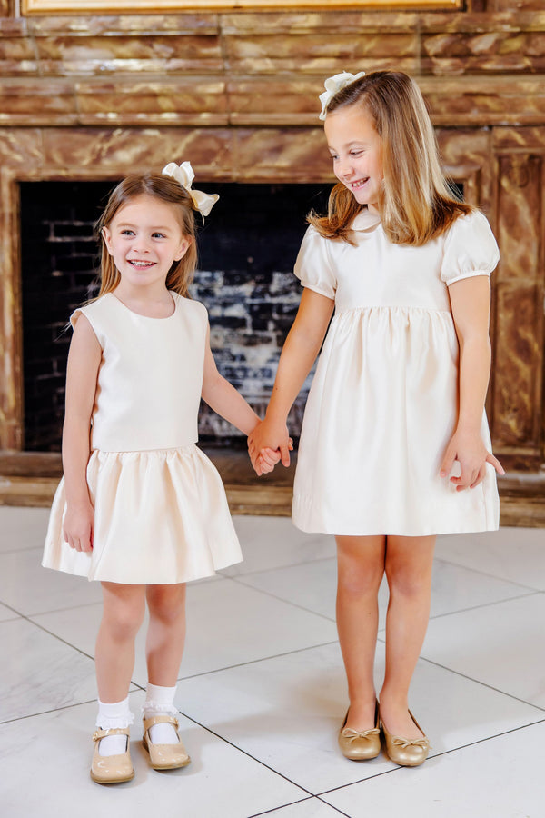 Two young girls in matching The Beaufort Bonnet Company Grandccasion™ Tessa Tops in Grand Isle Ivory Taffeta and coordinating hair bows smile and hold hands while standing indoors in front of a fireplace.