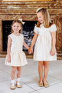 Two young girls in matching The Beaufort Bonnet Company Grandccasion™ Tessa Tops in Grand Isle Ivory Taffeta and coordinating hair bows smile and hold hands while standing indoors in front of a fireplace.