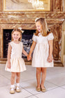 Two young girls dressed in The Beaufort Bonnet Company’s Grandccasion™ Cindy Lou Sash Dress in Grand Isle Ivory hold hands and smile while standing in front of a fireplace topped with a gold-framed mirror.