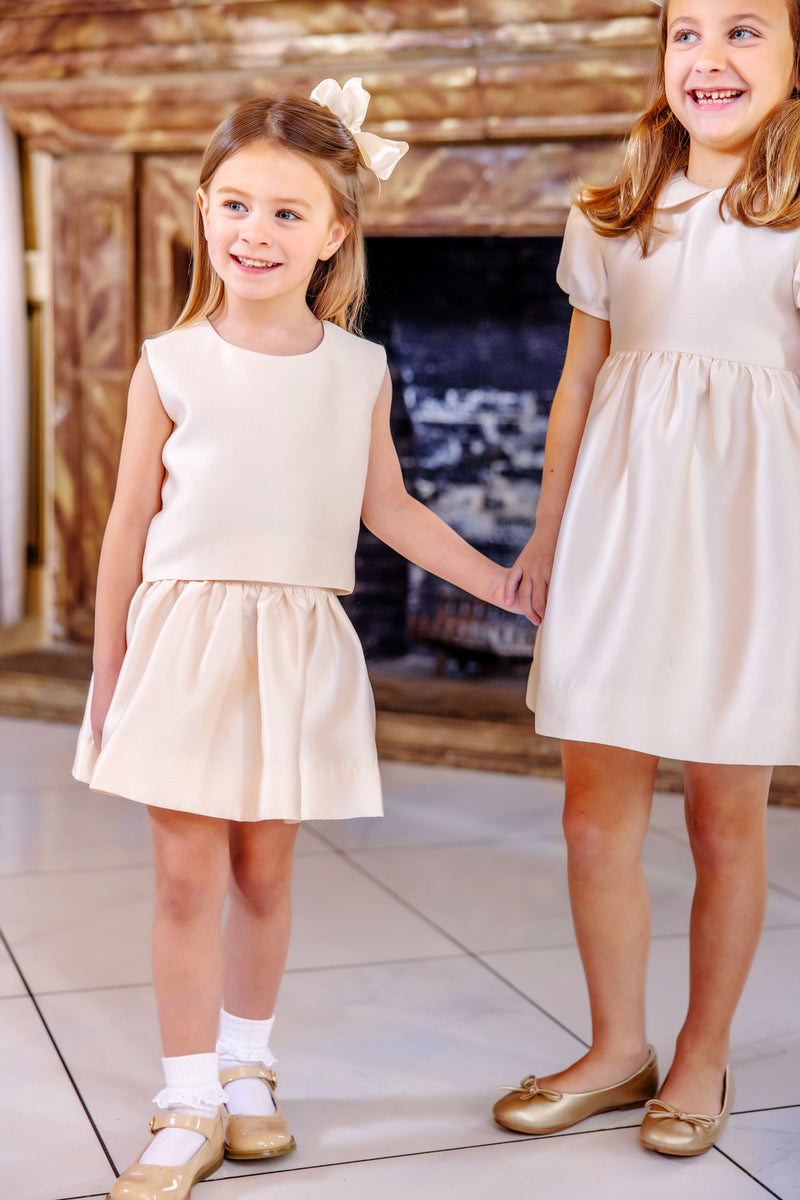Two young girls wearing the Grandccasion™ Tessa Top in Grand Isle Ivory Taffeta by The Beaufort Bonnet Company and gold shoes stand indoors, holding hands and smiling. A decorative fireplace in the background highlights their roles as flower girls.
