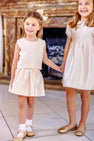 Two young girls wearing the Grandccasion™ Tessa Top in Grand Isle Ivory Taffeta by The Beaufort Bonnet Company and gold shoes stand indoors, holding hands and smiling. A decorative fireplace in the background highlights their roles as flower girls.