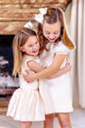 Two young girls in matching Grandccasion™ Cindy Lou Sash Dresses in Grand Isle Ivory by The Beaufort Bonnet Company, accessorized with hair bows, hug and smile while standing indoors in front of a fireplace.