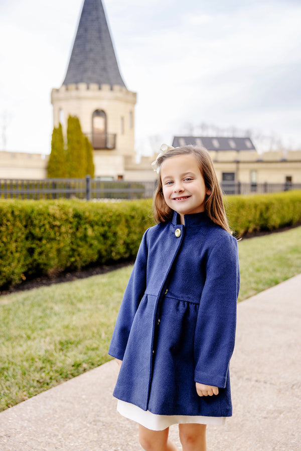 A young girl wearing The Beaufort Bonnet Company’s Penelope Peacoat in Nantucket Navy stands smiling on a path, with manicured hedges and a stone building featuring a tall pointed tower in the background.