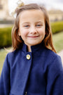 A young girl with light brown hair, adorned with a white bow, smiles at the camera while wearing The Beaufort Bonnet Company’s Penelope Peacoat in Nantucket Navy. She stands outdoors surrounded by greenery, with blurred buildings in the background.