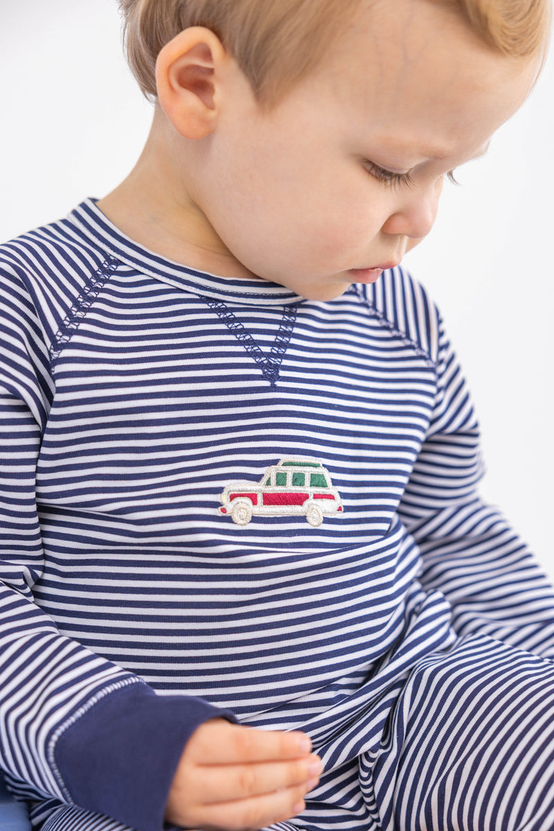 A young child in The Beaufort Bonnet Company’s Cassidy Comfy Crewneck Romper—Nantucket Navy Stripe with Wagon Embroidery—looks down, showing a side profile against a plain background.