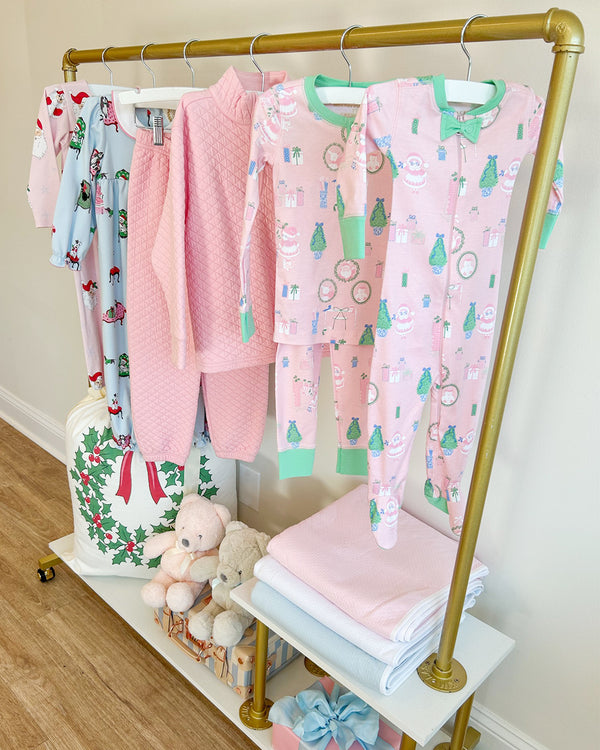 A gold clothes rack displays The Beaufort Bonnet Company’s Sara Jane’s Sweet Dream Set in the Mrs. Claus Collage with Grace Bay Green print—adorable children’s holiday pajamas adorned with festive patterns. Below, neatly folded blankets, a cuddly lamb plush, and a cheerful Christmas bag add to the cozy, festive scene.