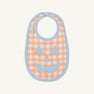 The Beaufort Bonnet Company’s Burp Me Bib - Seashore Sherbet Gingham & Beale Street Blue features an orange and white checkered pattern with light blue trim, making it a perfect fall baby essential. The front is adorned with a charming light blue jack-o-lantern face appliqué.