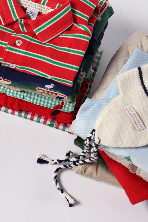 A neatly folded stack of brightly colored children’s clothes—including striped shirts, patterned sweaters such as The Beaufort Bonnet Company’s Hayden Half-Zip Sweater in Richmond Red with Nantucket Navy Stork, and a knit hat with navy and white braided ties—arranged on a white surface.