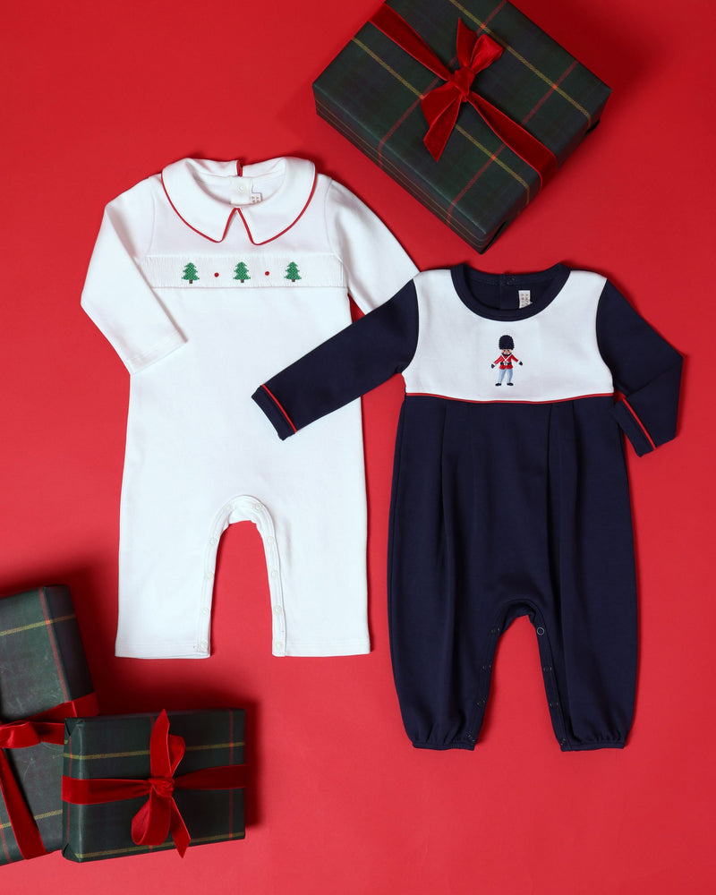 Two Samuel Smocked Romper - Worth Avenue White & Richmond Red with Trees outfits on a red background, one white with a smocked Christmas tree pattern and one navy and white toy soldier romper, surrounded by three wrapped gifts.