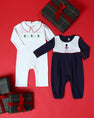 Two Samuel Smocked Romper - Worth Avenue White & Richmond Red with Trees outfits on a red background, one white with a smocked Christmas tree pattern and one navy and white toy soldier romper, surrounded by three wrapped gifts.