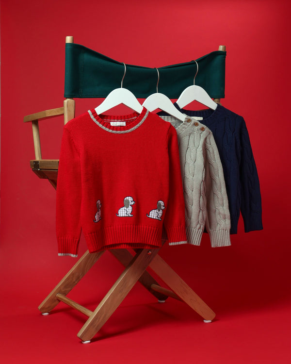Three children’s sweaters hanging on a director’s chair: an Isaac’s Intarsia Sweater - Richmond Red with Grantley Gray and Dogs, a gray cotton sweater with shoulder buttons, and a navy blue sweater.