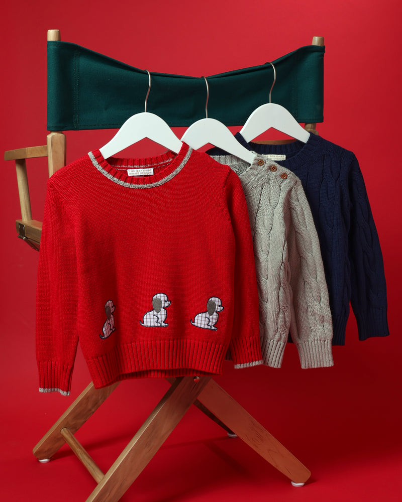 Three sweaters hanging on a director’s chair: a red Crawford Crewneck embroidered with dachshunds, a Crawford Crewneck - Grantley Gray with Brass Buttons cable-knit sweater, and a solid navy cable-knit sweater.