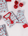 The Sutton’s Sweet Dream Set - Santa’s Laundry Line with Richmond Red from The Beaufort Bonnet Company, featuring light blue Pima cotton pajamas with red trim and a festive holiday print, is folded on a white surface beside three shiny red Christmas ornaments.