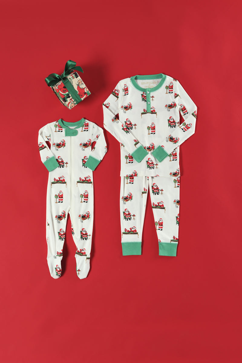 Two Sutton’s Sweet Dream Sets in The Beaufort Bonnet Company’s Serious Santa Business print—one footed and one not—are laid out on a red background next to a gift wrapped with a green ribbon.