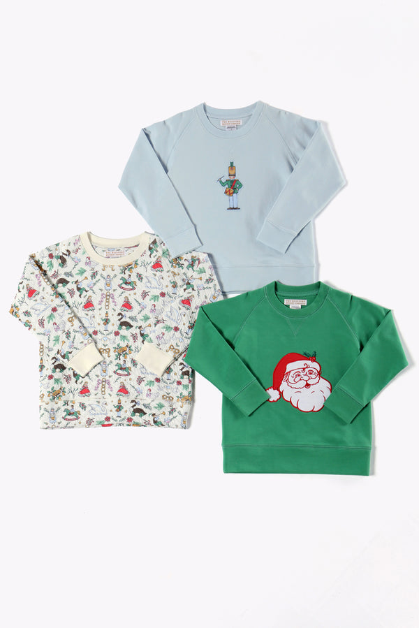 Three children’s sweatshirts from The Beaufort Bonnet Company are laid flat on a white background: one pale blue “Cassidy Comfy Crewneck - 12 Days of Christmas” featuring a nutcracker graphic, one green quilted crewneck with a Santa face, and one cream holiday party top with a colorful holiday-themed pattern.