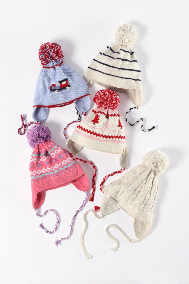 Five colorful winter hats with pom-poms and ear flaps are laid out on a white surface. Each hat showcases a unique pattern, featuring stripes, geometric designs, and festive motifs. Among them is the playful Parrish Pom Pom Hat - Palmetto Pearl with Bows & Trees from The Beaufort Bonnet Company, adding an extra touch of cheer to the collection.