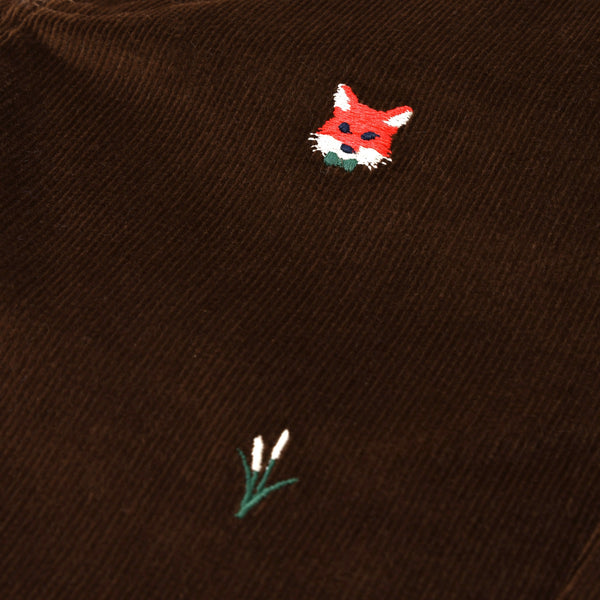 A close-up of The Beaufort Bonnet Company’s Critter Prep School Pants in Chelsea Chocolate Corduroy highlights an embroidered red fox head alongside a simple green plant with two white buds.