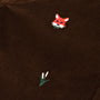 A close-up of The Beaufort Bonnet Company’s Critter Prep School Pants in Chelsea Chocolate Corduroy highlights an embroidered red fox head alongside a simple green plant with two white buds.
