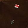 A close-up of The Beaufort Bonnet Company’s Critter Prep School Pants in Chelsea Chocolate Corduroy highlights an embroidered red fox head alongside a simple green plant with two white buds.