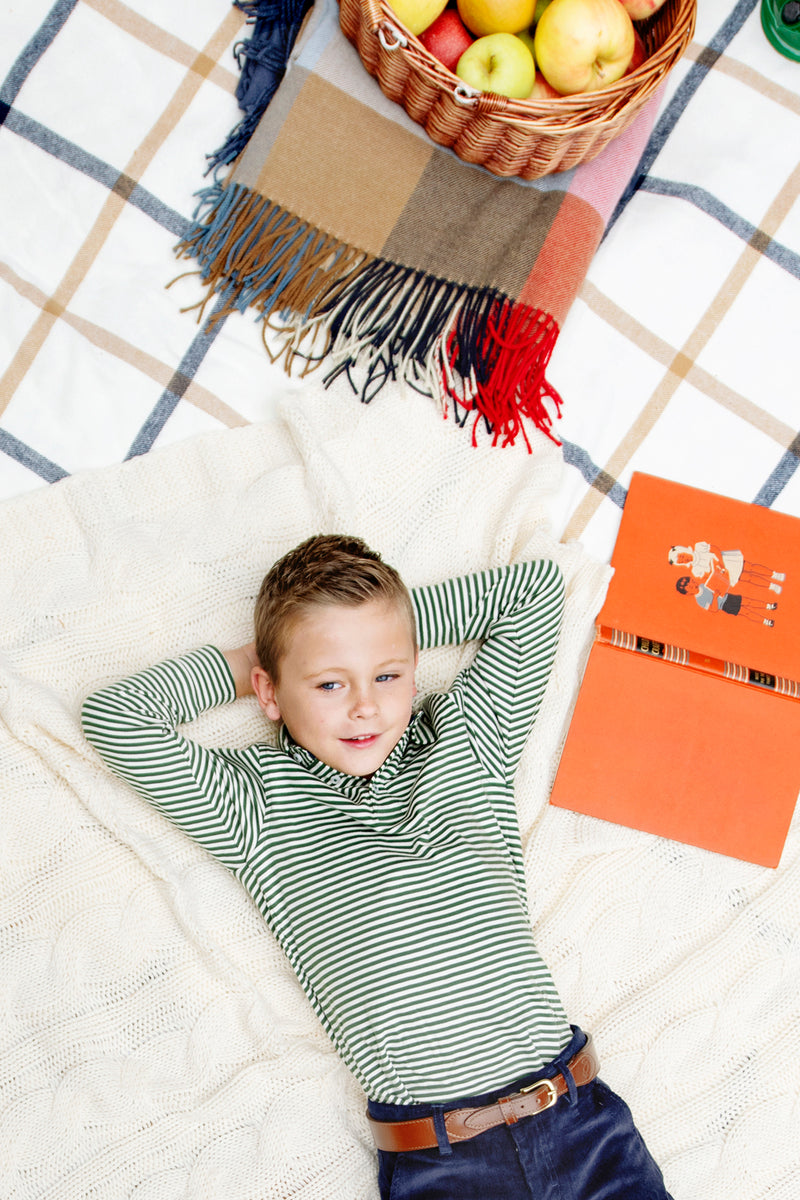 A young boy reclines on a blanket outdoors, smiling with his hands behind his head. Next to him rest a plaid blanket, a basket of apples, and an open orange book. He wears The Beaufort Bonnet Company’s Long Sleeve Prim & Proper Polo & Onesie in Grier Green & Palmetto Pearl Stripe with Nantucket Navy Stork, adding to the preppy charm of the scene.