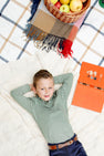 A young boy reclines on a blanket outdoors, smiling with his hands behind his head. Next to him rest a plaid blanket, a basket of apples, and an open orange book. He wears The Beaufort Bonnet Company’s Long Sleeve Prim & Proper Polo & Onesie in Grier Green & Palmetto Pearl Stripe with Nantucket Navy Stork, adding to the preppy charm of the scene.