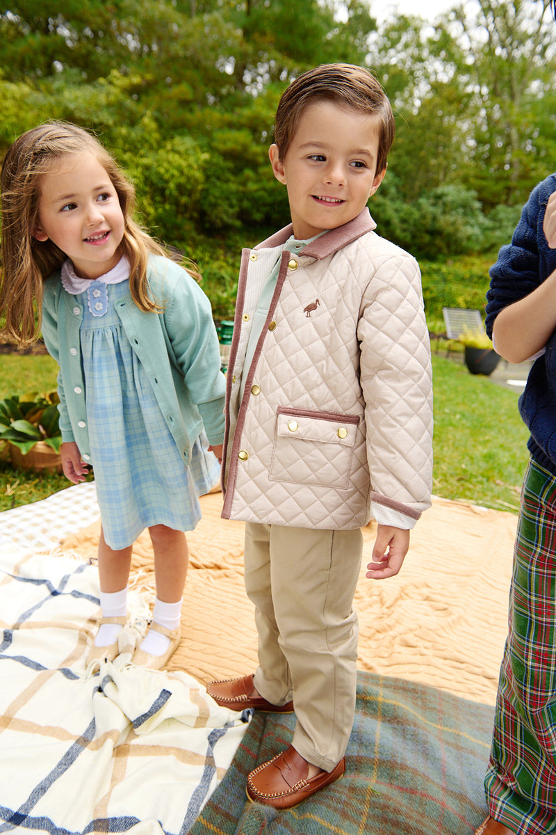 Two young children stand on a picnic blanket outdoors. The girl wears a blue dress and light green cardigan, while the boy is dressed in The Beaufort Bonnet Company’s Caldwell Quilted Coat in Keeneland Khaki with a Gray Bay Brown Corduroy collar and khaki pants. Both are smiling on a grassy area with trees in the background.
