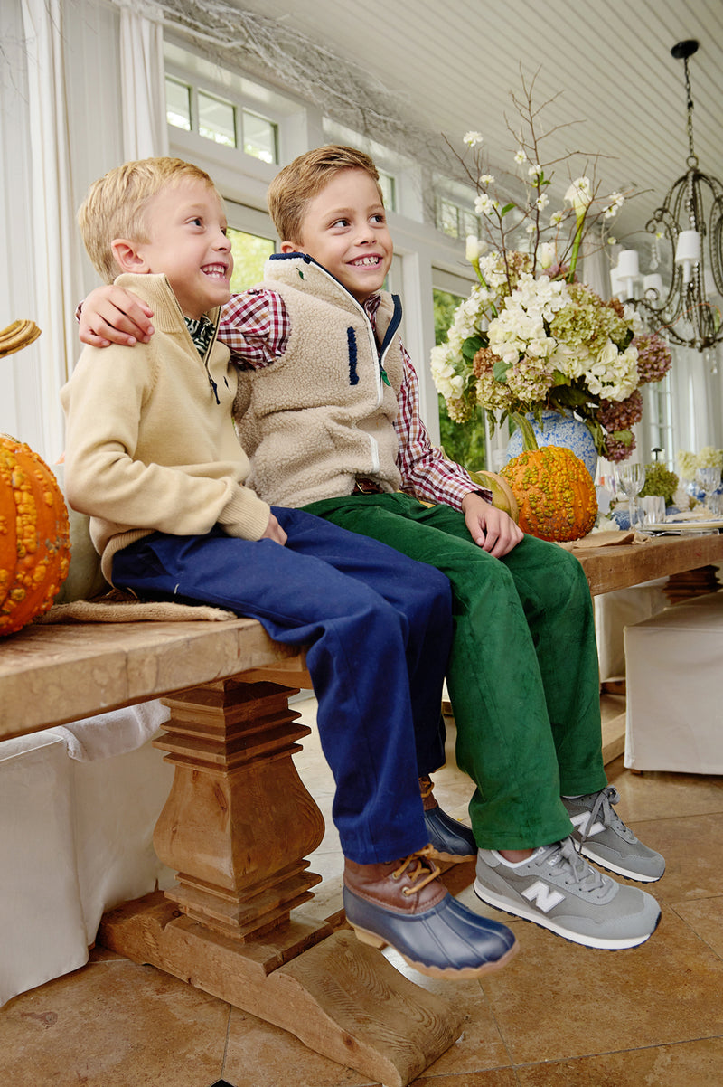 Two young boys sit smiling on a wooden bench indoors, surrounded by autumn decorations like pumpkins and flowers. One is cozily layered in The Beaufort Bonnet Company’s Van Camp Vest in Keeneland Khaki Sherpa with Grier Green, making it an ideal fall piece. Both boys wear colorful pants and sneakers, looking happy and relaxed together.