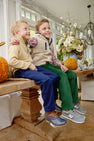 Two young boys sit smiling on a wooden bench indoors, surrounded by autumn decorations like pumpkins and flowers. One is cozily layered in The Beaufort Bonnet Company’s Van Camp Vest in Keeneland Khaki Sherpa with Grier Green, making it an ideal fall piece. Both boys wear colorful pants and sneakers, looking happy and relaxed together.