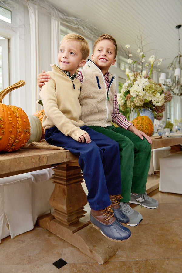 Two young boys sit on a wooden table adorned with pumpkins and flowers, smiling with their arms around each other. Both wear fleece jackets and corduroy pants in a bright, sunlit room—one sporting The Beaufort Bonnet Company’s Dean’s List Dress Shirt in Easton Pond Plaid beneath his jacket for a classic touch.