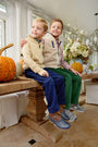 Two young boys sit on a wooden table adorned with pumpkins and flowers, smiling with their arms around each other. Both wear fleece jackets and corduroy pants in a bright, sunlit room—one sporting The Beaufort Bonnet Company’s Dean’s List Dress Shirt in Easton Pond Plaid beneath his jacket for a classic touch.