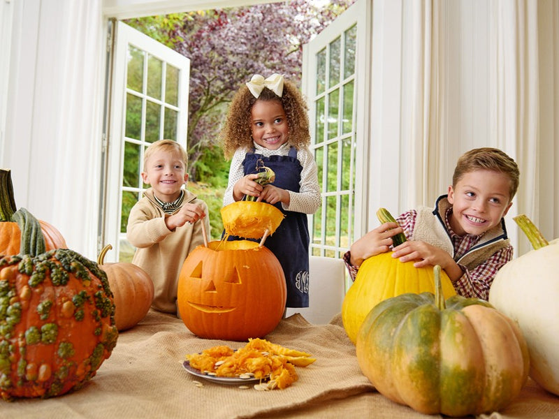 Three smiling children carve pumpkins indoors, surrounded by large pumpkins and pumpkin seeds on the table. One girl wears The Beaufort Bonnet Company’s Tatum’s Turtleneck & Onesie in Nantucket Navy Microdot—crafted from soft pima cotton—as sunlight streams through open French doors, revealing trees outside.