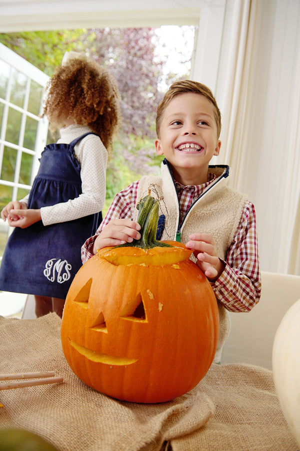 A smiling boy sits at a table with a carved pumpkin, wearing The Beaufort Bonnet Company’s Van Camp Vest in Keeneland Khaki Sherpa with Grier Green over a plaid shirt. A girl in a blue dress stands nearby, turned away, as sunlight streams in through large windows behind them.