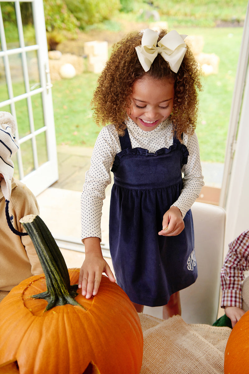 A young girl with curly hair and a large white bow smiles as she touches a pumpkin on an indoor table. She is wearing The Beaufort Bonnet Company’s Tatum’s Turtleneck & Onesie in Nantucket Navy Microdot. An open door to the garden is visible behind her.