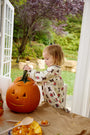 A young child wearing The Beaufort Bonnet Company’s Long Sleeve Banks Bow Dress in the Happy Hunting with Palmetto Pearl & Nantucket Navy pattern scoops out a large jack-o-lantern pumpkin at a table near open doors, with autumn scenery and hay bales visible outside. The cozy scene highlights the dress’s soft comfort, perfect for fall festivities.