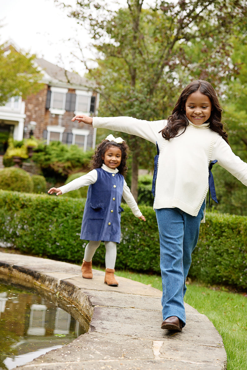 Two young girls walk playfully along the edge of a garden pond, arms outstretched for balance. Dressed in cozy pieces from their girls autumn wardrobe—including The Beaufort Bonnet Company’s Shae Sweater Vest in Palmetto Pearl with Nantucket Navy, crafted from 100% cotton sweater material—they explore together as a house and lush greenery appear in the background.