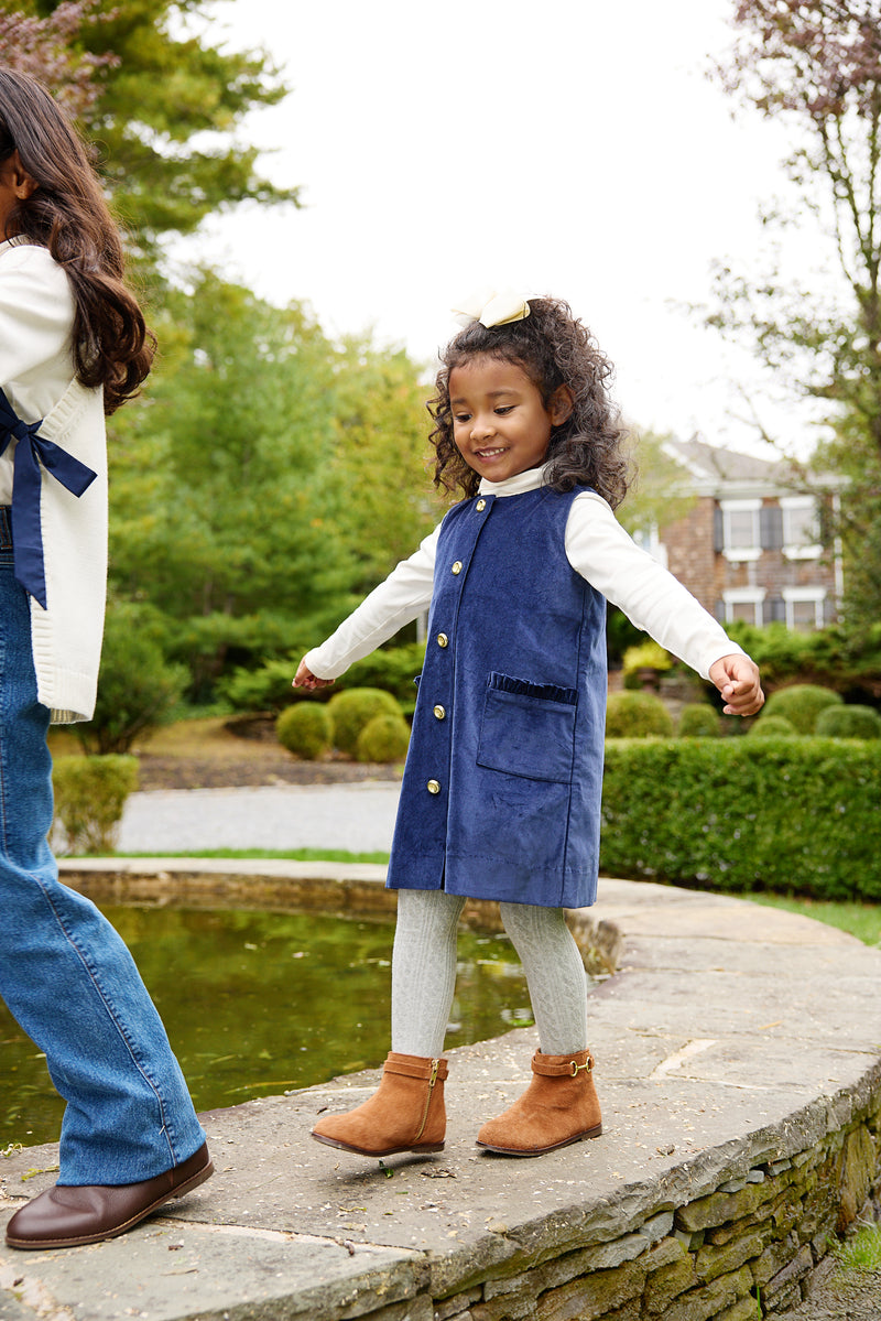 A young girl in a blue corduroy Juliet Jumper and gray Jefferies Cable Tights - Gray Heather walks along a stone ledge near a garden pond.