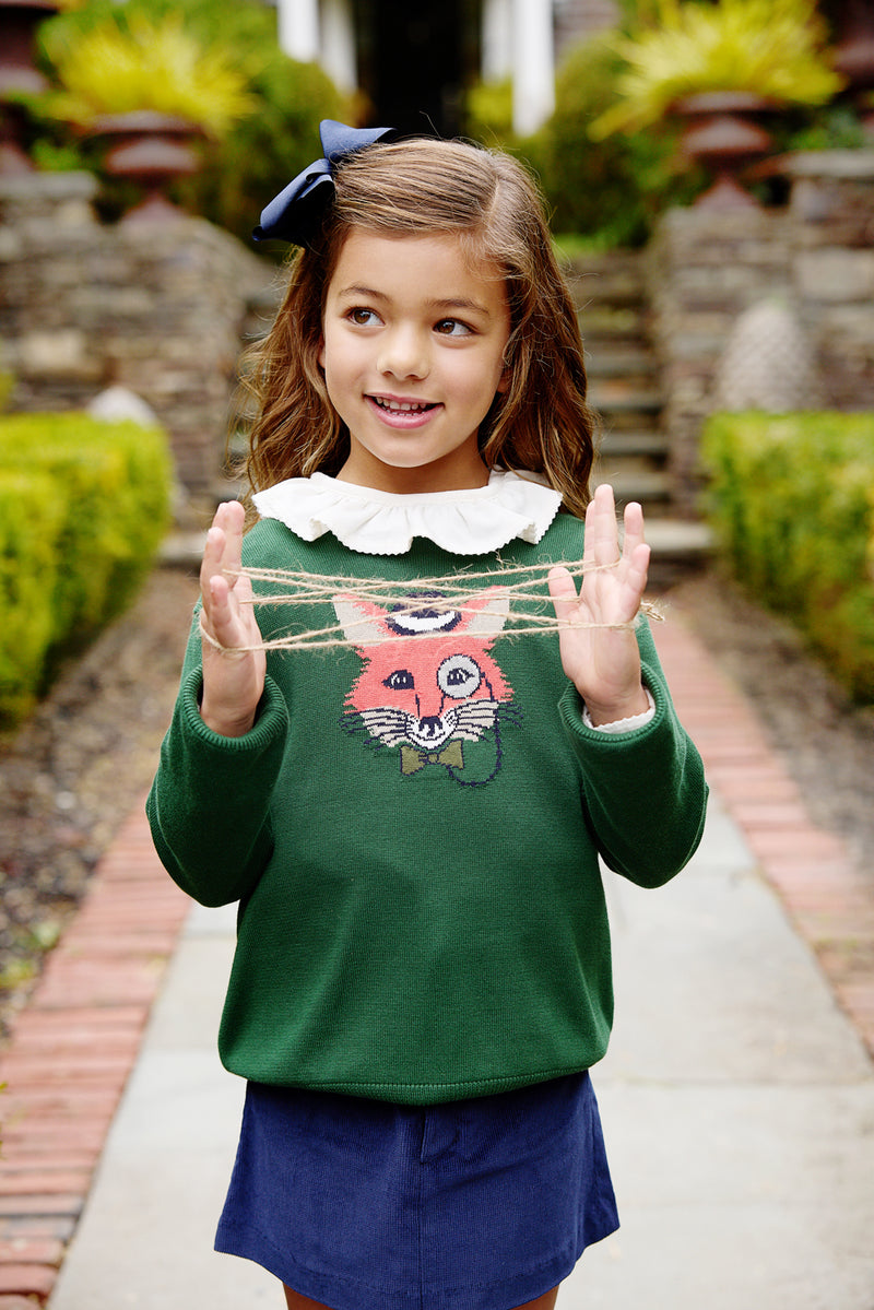 A young girl wearing The Beaufort Bonnet Company’s Isaac’s Intarsia Sweater in Grier Green with Fox, featuring a charming white collar, plays cat’s cradle with string on a garden path.