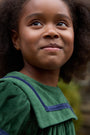 A young girl with curly hair smiles softly, wearing The Beaufort Bonnet Company’s Short Sleeve Patsy’s Party Dress in Grier Green Velveteen with Nantucket Navy trim. Her charming holiday dress stands out as she poses outdoors, surrounded by lush greenery.