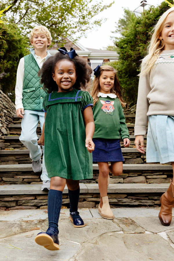 Four smiling kids in autumn outfits walk down outdoor stone steps. The girl in front wears The Beaufort Bonnet Company’s Isaac’s Intarsia Sweater in Grier Green with Fox, while the others are dressed in cozy cotton sweaters and skirts. Trees and a house create a charming background.