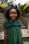 A young girl with curly hair, wearing The Beaufort Bonnet Company’s Short Sleeve Patsy’s Party Dress in Grier Green Velveteen with Nantucket Navy trim and bow, smiles outdoors in front of stone steps and greenery—a perfect choice for the holidays.
