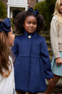A young girl wearing The Beaufort Bonnet Company’s Penelope Peacoat in Nantucket Navy, featuring gold buttons, stands outdoors holding hands with others. She is smiling, has a navy hair bow, and is accompanied by another girl and an adult.