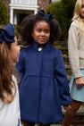 A young girl wearing The Beaufort Bonnet Company’s Penelope Peacoat in Nantucket Navy, featuring gold buttons, stands outdoors holding hands with others. She is smiling, has a navy hair bow, and is accompanied by another girl and an adult.