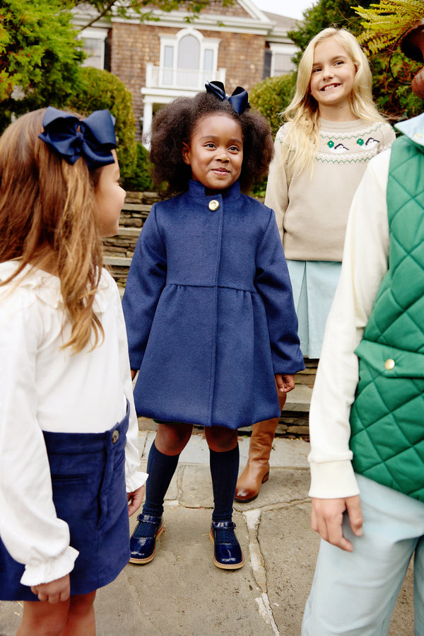 Four children stand outside, smiling. The girl in the center wears The Beaufort Bonnet Company’s Penelope Peacoat in Nantucket Navy with a matching bow, flanked by another girl dressed in navy and white, a blonde girl in a light sweater, and a child wearing a green quilted vest. Behind them are stone steps and a house.